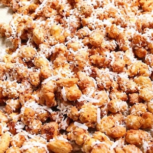May include: A collection of roasted peanuts coated in a golden-brown caramelized sugar glaze and sprinkled with shredded white coconut flakes. The texture appears crunchy and uneven, with the nuts clustered together in a dense, textured mixture.