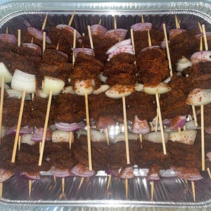 May include: A silver foil tray filled with meat skewers. Each skewer has pieces of seasoned meat, red onion, and white onion. The meat is covered in a dark brown spice mix, ready for cooking or serving.
