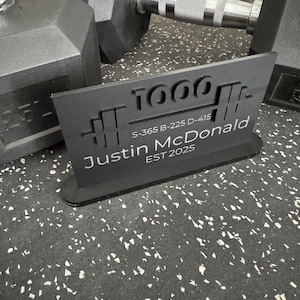 May include: A black sign with the text "1000" and a barbell graphic. The sign also includes the name "Justin McDonald" and the establishment year "EST 2025". Dumbbells are in the background.