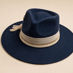 May include: A navy blue fedora hat with a wide brim. The hat features a cream-colored band with gold-colored chain and white bead accents. A cream-colored ribbon is attached to the side of the hat. The hat is a stylish accessory.