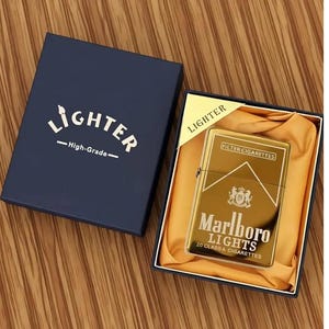 May include: A gold-coloured Marlboro Lights lighter in a navy blue box. The lighter has the Marlboro logo and text. The box is labelled "Lighter - High-Grade-". The lighter is in a gold satin-like material.