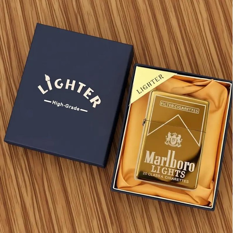May include: A gold-coloured Marlboro Lights lighter in a navy blue box. The lighter has the Marlboro logo and text. The box is labelled "Lighter - High-Grade-". The lighter is in a gold satin-like material.