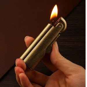 Peut inclure: Un briquet en laiton de style vintage avec une flamme allum&eacute;e. Le briquet est tenu dans une main, mettant en valeur sa finition m&eacute;tallique et son design. La flamme est orange vif et jaune, sur un fond plus sombre.