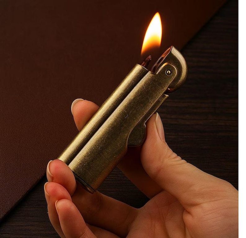 Peut inclure: Un briquet en laiton de style vintage avec une flamme allum&eacute;e. Le briquet est tenu dans une main, mettant en valeur sa finition m&eacute;tallique et son design. La flamme est orange vif et jaune, sur un fond plus sombre.