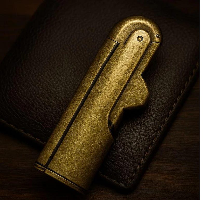 Peut inclure: Un briquet de couleur laiton antique avec un corps rectangulaire et un dessus arrondi. Le briquet a un couvercle &agrave; charni&egrave;re et un design incurv&eacute; et ergonomique. La surface montre des signes d'usure, lui donnant un aspect vintage. Le briquet est pos&eacute; sur une surface en cuir brun fonc&eacute;.