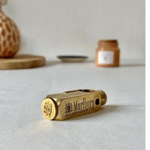 May include: A gold-colored cigarette case with the Marlboro logo and text. The case is rectangular with rounded edges and a small, cylindrical knob. The text reads "Marlboro 20 CLASS A CIGARETTES".