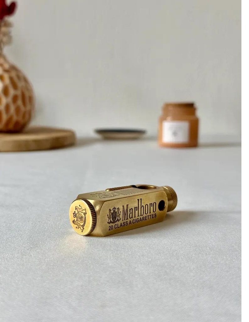 May include: A gold-colored cigarette case with the Marlboro logo and text. The case is rectangular with rounded edges and a small, cylindrical knob. The text reads "Marlboro 20 CLASS A CIGARETTES".
