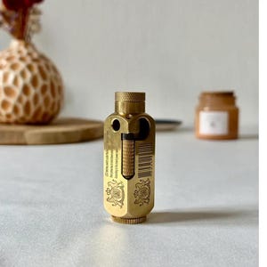 May include: A brass-colored, vintage-style lighter with a textured top and intricate engravings. The lighter has a rectangular body with a hinged section and a barcode. The background includes a vase with flowers and a small jar.