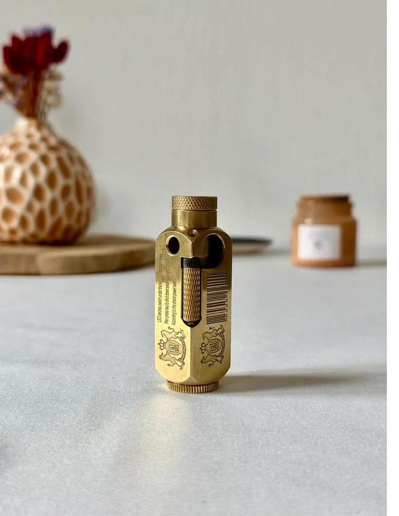 May include: A brass-colored, vintage-style lighter with a textured top and intricate engravings. The lighter has a rectangular body with a hinged section and a barcode. The background includes a vase with flowers and a small jar.