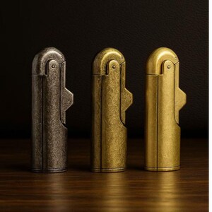 Peut inclure: Trois briquets de style vintage align&eacute;s, chacun avec une finition unique. Le premier est en bronze fonc&eacute; vieilli, le second en laiton chaud et le troisi&egrave;me en or brillant. Les briquets ont un corps rectangulaire avec un dessus incurv&eacute; et un levier lat&eacute;ral.