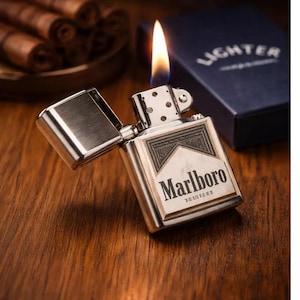 May include: A silver-coloured, open lighter with a flame, featuring the Marlboro logo. The lighter is on a wooden surface, with a box labelled "Lighter" in the background. Cigars are visible in the background.