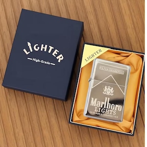 May include: A silver-coloured lighter with the text "Marlboro Lights" and a logo, presented in a navy blue box with the word "Lighter" printed on it. The lighter is in a black box with yellow satin fabric.