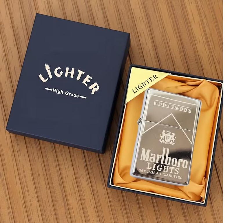 May include: A silver-coloured lighter with the text "Marlboro Lights" and a logo, presented in a navy blue box with the word "Lighter" printed on it. The lighter is in a black box with yellow satin fabric.