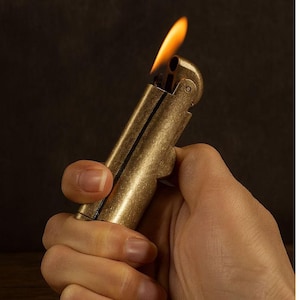 Peut inclure: Un briquet en laiton de style vintage avec une flamme allum&eacute;e. Le briquet est tenu dans une main, mettant en valeur sa finition m&eacute;tallique et son design. La flamme est orange vif et jaune sur un fond sombre.
