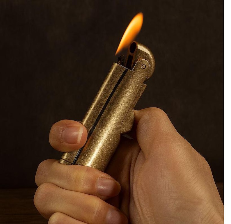 Peut inclure: Un briquet en laiton de style vintage avec une flamme allum&eacute;e. Le briquet est tenu dans une main, mettant en valeur sa finition m&eacute;tallique et son design. La flamme est orange vif et jaune sur un fond sombre.