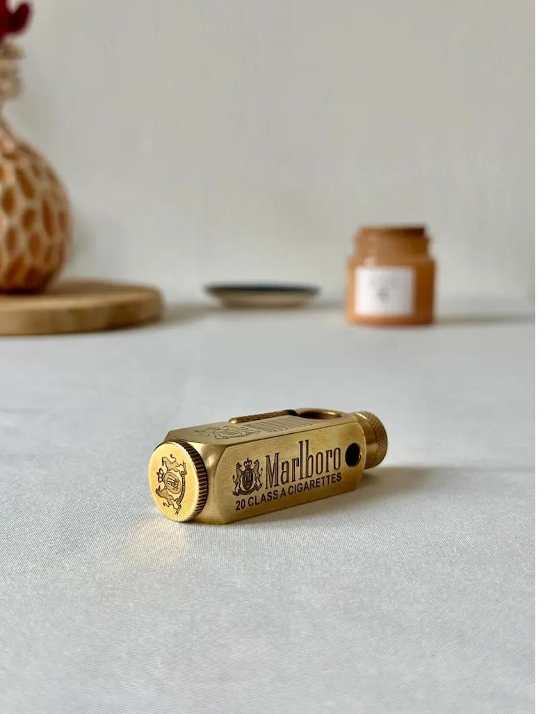 May include: A gold-colored lighter with the Marlboro logo and text. The lighter has a rectangular body with rounded edges and a textured surface. The text "20 CLASS A CIGARETTES" is also visible.