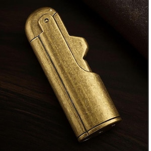 Peut inclure: Un briquet en laiton de style vintage avec un couvercle &agrave; charni&egrave;re et un corps rectangulaire. Le briquet a une finition antique us&eacute;e et une d&eacute;coupe d&eacute;corative sur le c&ocirc;t&eacute;. Le briquet est pr&eacute;sent&eacute; sur une surface en bois sombre.