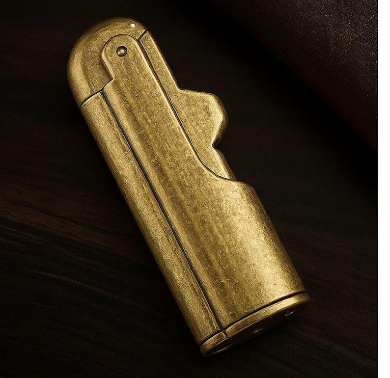 Peut inclure: Un briquet en laiton de style vintage avec un couvercle &agrave; charni&egrave;re et un corps rectangulaire. Le briquet a une finition antique us&eacute;e et une d&eacute;coupe d&eacute;corative sur le c&ocirc;t&eacute;. Le briquet est pr&eacute;sent&eacute; sur une surface en bois sombre.
