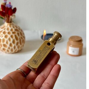 May include: A vintage brass lighter with the text "Marlboro 20 CLASS A CIGARETTES" engraved on the side. The lighter is cylindrical with a textured top and bottom. The lighter is held in a hand, with a lit candle in the background.
