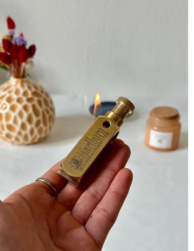 May include: A vintage brass lighter with the text "Marlboro 20 CLASS A CIGARETTES" engraved on the side. The lighter is cylindrical with a textured top and bottom. The lighter is held in a hand, with a lit candle in the background.