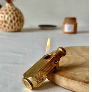 May include: A brass-colored lighter with the word "Marlboro" engraved on the side. The lighter is lit, with a flame visible. The lighter is resting on a wooden surface.