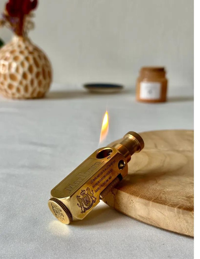 May include: A brass-colored lighter with the word "Marlboro" engraved on the side. The lighter is lit, with a flame visible. The lighter is resting on a wooden surface.