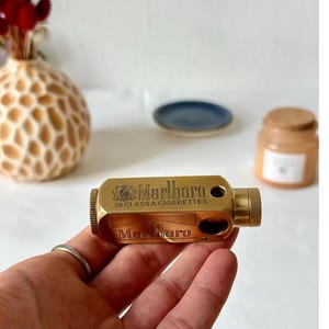 May include: A brass cigarette case with the Marlboro logo and the text "20 CLASS A CIGARETTES". The case is rectangular with a textured end and is held in a hand. A ring is visible on the finger.