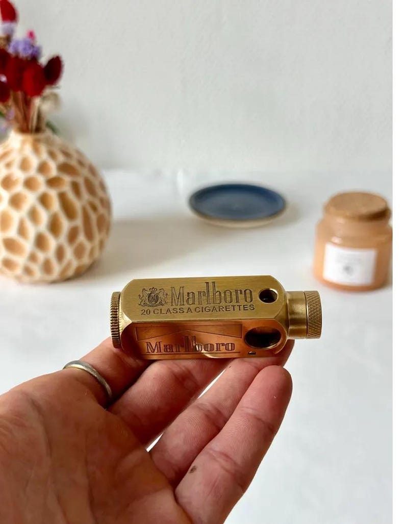 May include: A brass cigarette case with the Marlboro logo and the text "20 CLASS A CIGARETTES". The case is rectangular with a textured end and is held in a hand. A ring is visible on the finger.