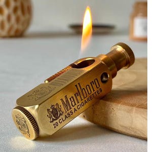 May include: A gold-colored lighter with the word "Marlboro" and "20 CLASS A CIGARETTES" printed on the side. The lighter has a flame lit, and a decorative emblem. The lighter is on a wooden surface.