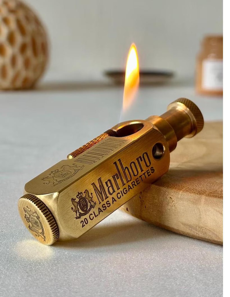 May include: A gold-colored lighter with the word "Marlboro" and "20 CLASS A CIGARETTES" printed on the side. The lighter has a flame lit, and a decorative emblem. The lighter is on a wooden surface.