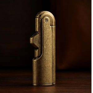 Peut inclure: Un briquet en laiton de style vintage avec un design unique. Le corps rectangulaire a un dessus arrondi et une surface textur&eacute;e. La finition en laiton lui donne un aspect antique. Le briquet est con&ccedil;u pour &ecirc;tre tenu &agrave; la main.