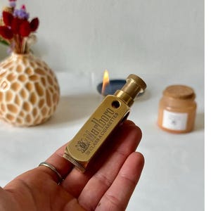 May include: A vintage brass lighter with the Marlboro logo and text. The lighter is rectangular with a textured end and a knurled top. The lighter is held in a hand, with a lit candle and a decorative vase in the background.