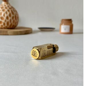 May include: A gold-colored, rectangular lighter with ornate crest detailing on the end. The lighter has a textured surface and a hinged top. The background includes a decorative vase and a small jar.