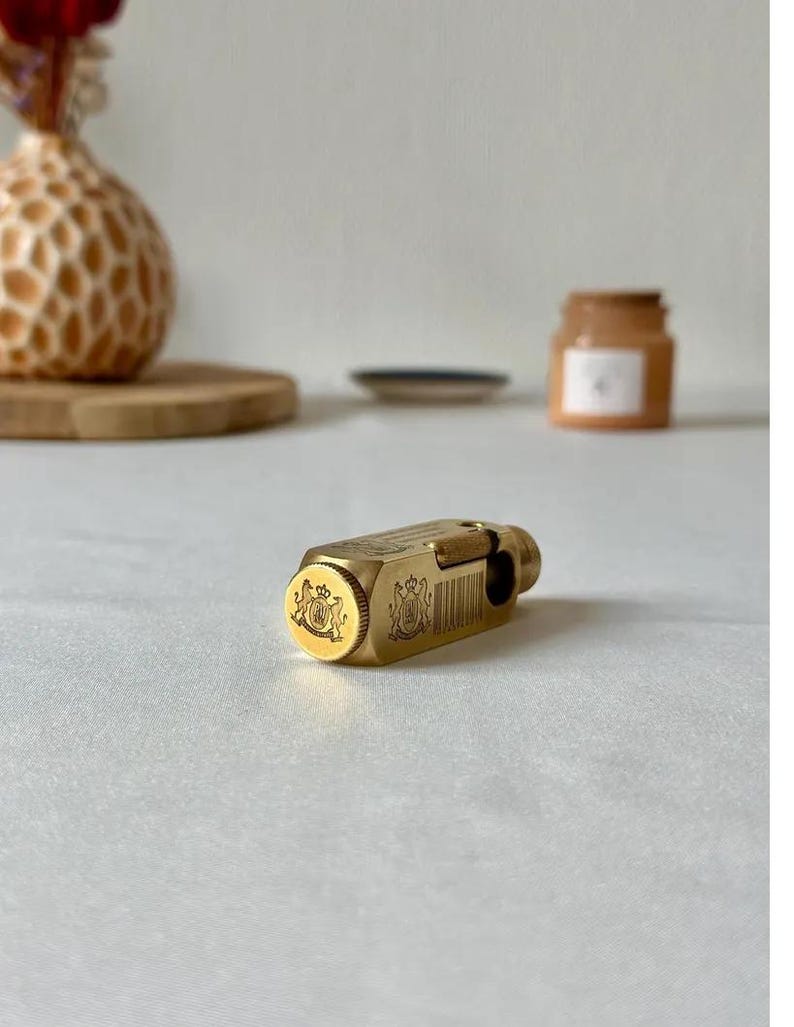 May include: A gold-colored, rectangular lighter with ornate crest detailing on the end. The lighter has a textured surface and a hinged top. The background includes a decorative vase and a small jar.