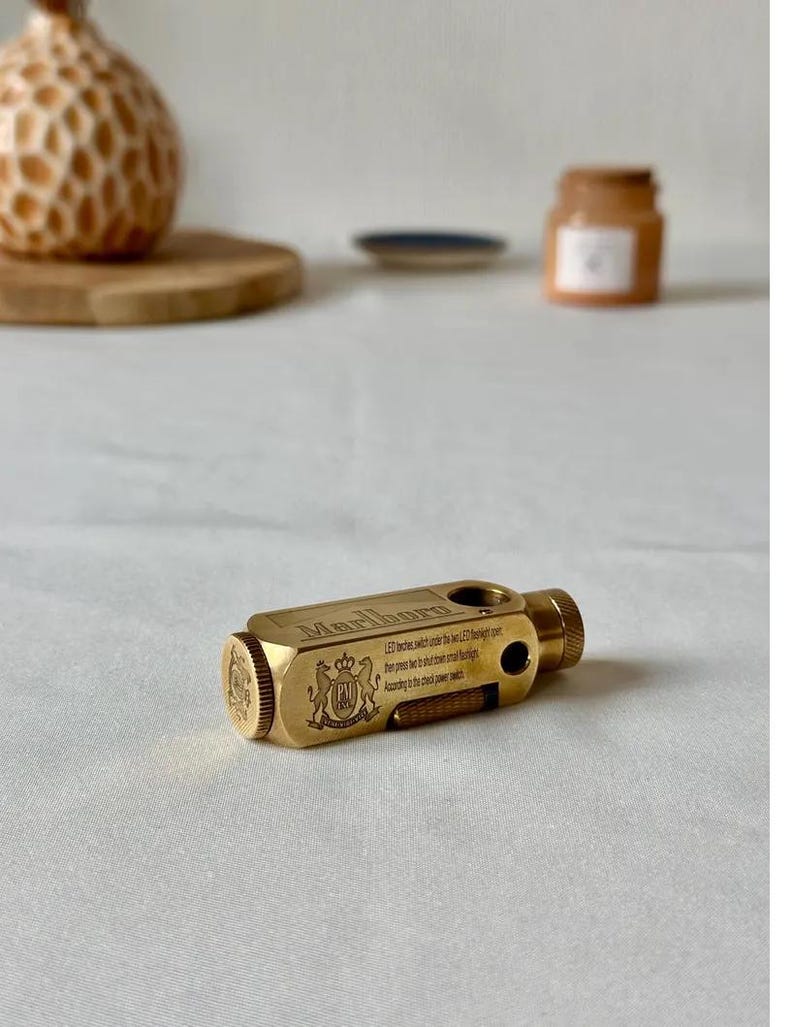 May include: A brass-colored, rectangular lighter with the word "Marlboro" engraved on the top. The lighter has a textured end and a small switch. The text "LED lights..." is also visible. A decorative crest is on the side.