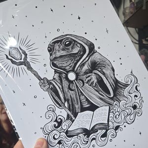 May include: Black and white illustration of a wizard toad holding a staff with a crystal, wearing a hooded robe, and standing in front of an open book. The artwork features swirling lines and stars, creating a mystical atmosphere.