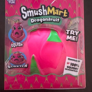 Peut inclure: Un jouet SmushMart Dragonfruit dans une boîte rose avec des accents verts. L'emballage porte les textes SmushMart, Dragonfruit, Try Me, Squish, Stretch et Grocery List 100% Squishy Dragonfruit. Le jouet est un objet rond, rose vif et mou.
