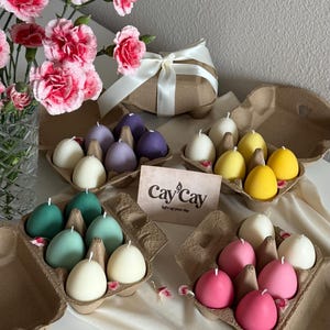 May include: Egg-shaped candles in various colors, including white, purple, yellow, green, and pink, are displayed in cardboard egg cartons. A card with the text "Cay Cay light up your day" is visible. A bouquet of pink flowers and a wrapped gift are also present.