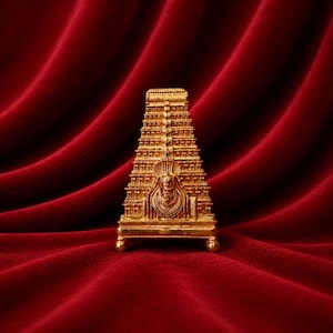 May include: A gold-coloured miniature replica of a temple gopuram tower featuring intricate architectural carvings, multiple tiered levels, and a central deity figure, standing on a small rectangular base with four rounded feet.