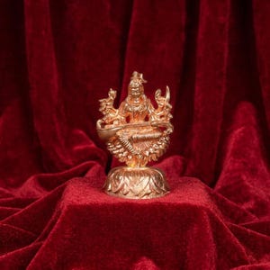 May include: A metallic gold-toned figurine depicting a seated deity with four arms, holding a stringed instrument across the lap. The figure is positioned on a base shaped like an open lotus flower with detailed petals and a polished finish.