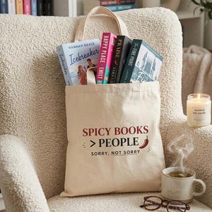 Peut inclure: Un sac fourre-tout en toile crème rempli de livres, avec le texte "SPICY BOOKS > PEOPLE, SORRY, NOT SORRY." Une bougie allumée, une tasse de thé et des lunettes sont à côté du sac. Les livres incluent "Icebreaker" et "Happy Place."