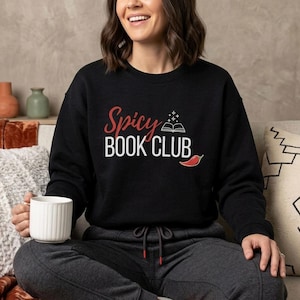 Peut inclure: Sweat-shirt noir avec l'inscription "Spicy Book Club" en rouge et blanc, avec un livre ouvert et un piment rouge. La personne tient une tasse blanche.