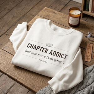 Peut inclure: Un sweat-shirt blanc avec l'inscription "CHAPTER ADDICT" et la phrase "just one more (I'm lying.)" imprimée en noir. Le sweat-shirt est posé sur une surface en bois, avec une bougie allumée et un marque-page.