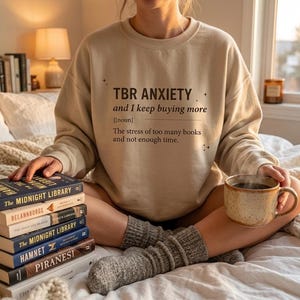 Peut inclure: Un sweat-shirt beige avec le texte "TBR ANXIETY and I keep buying more" et une définition. Une pile de livres est à côté de la personne portant le sweat-shirt. Une tasse de café est posée sur les genoux de la personne.