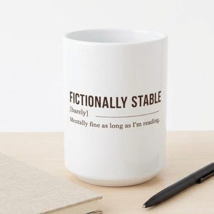 Peut inclure: Un mug en céramique blanc avec le texte "FICTIONALLY STABLE" en marron, avec la définition "Mentally fine as long as I'm reading." Un stylo noir et un livre sont sur la table à côté du mug.