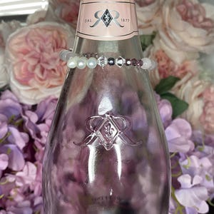 May include: A pink Ruffino sparkling wine bottle with a decorative bracelet. The bottle has a pink label with the year 1877. The bracelet is made of white pearls, gray beads, and purple crystals. The background features pink and purple flowers.