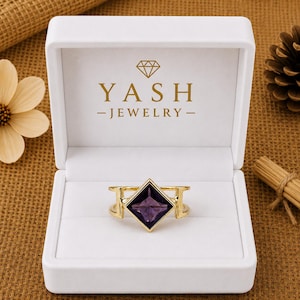 May include: A gold ring featuring a square-cut purple gemstone set in a white jewelry box with the text YASH JEWELRY and a diamond icon printed on the inside lid. The ring has a split band design and is centered within the white velvet interior.