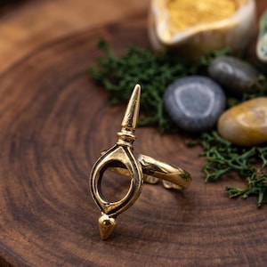 May include: A gold-toned metal ring featuring a central vertical design with a pointed top and bottom, surrounding an open circular center. The band is smooth and polished, showcasing a metallic finish with subtle dark accents in the recessed areas.