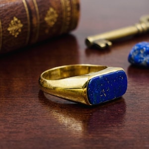 Puede incluir: Un anillo de sello de oro con una incrustación rectangular de piedra lapis lázuli azul intenso con motas doradas naturales. El anillo presenta una banda de oro pulida y lisa que se estrecha ligeramente hacia la parte posterior.