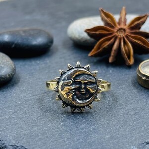 May include: A gold-toned metal ring featuring a circular centerpiece with a raised, detailed relief of a sun and crescent moon face. The design includes pointed rays extending from the perimeter, with a dark, oxidized finish in the recessed areas.
